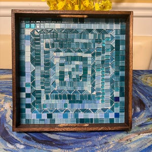 May include: A square wooden serving tray featuring a mosaic inlay of glass tiles in various shades of blue, teal, and turquoise. The tiles are arranged in a concentric square pattern with a mix of square and triangular shapes throughout the design.
