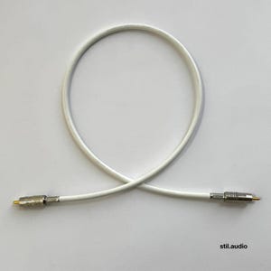 May include: A white audio cable looped into a circle with silver-toned metal connectors at each end. The connectors feature a textured grip and a gold-colored center pin. The text stil.audio is printed in the bottom right corner of the image.