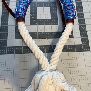 May include: A white rope halter with a blue and red braided top section. The halter features brown leather accents and a long, white tassel at the bottom. The rope is thick and appears durable.