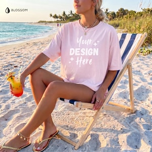May include: A light pink t-shirt with the text "Your DESIGN Here" is worn by a person sitting in a beach chair. The person is holding a tropical drink with a straw. The beach chair has blue and white stripes. Gold sandals are worn.