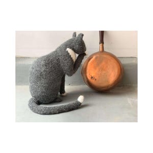 Puede incluir: Un gato gris tejido a mano con marcas blancas, sentado junto a una sartén de cobre con un mango de madera. El gato se está lavando la cara. La sartén es redonda y tiene un mango marrón oscuro.