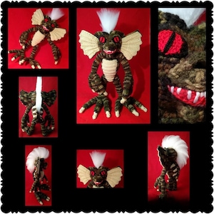 May include: A crocheted creature doll featuring a brown and green textured body, large cream-colored ears, a white faux fur mohawk, and red eyes with black pupils. The figure has long arms, three-toed feet, and a wide mouth with white teeth.