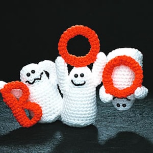 May include: Three handmade crochet ghosts spelling "BOO" with orange rings. The ghosts are white with black embroidered eyes and mouths. The background is a solid black.