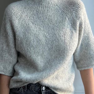 May include: A pale grey knitted sweater with short sleeves and a high neckline. The raglan sleeve design is visible. The fabric has a soft, textured appearance. The neutral colour of the sweater makes it versatile for various outfits.