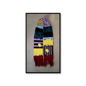 Op de afbeelding: Een kleurrijke sjaal in patchwork-stijl met een gefranjerde onderkant. De sjaal heeft een verscheidenheid aan kleuren en patronen, waaronder geel, zwart, blauw, paars en rood. De sjaal is handgemaakt en lijkt gebreid of gehaakt te zijn.