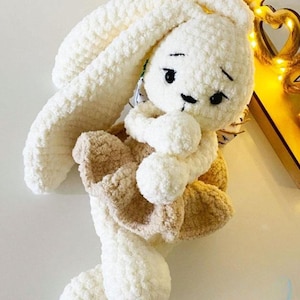 Könnte beinhalten: Ein gehäkeltes Plüschhasenspielzeug aus weichem, weißem Garn mit langen Ohren und einem beigen Rock. Der Hase hat gestickte schwarze Augen und eine kleine schwarze Nase. Die Garnstruktur ist dick und flauschig, was dem Stofftier ein rundes Aussehen verleiht.