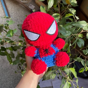 May include: A handmade plush Spider-Man doll. The doll features a red head and limbs, with white eye shapes and a blue torso. A black spider emblem is on the chest. The doll is made of soft, textured yarn.