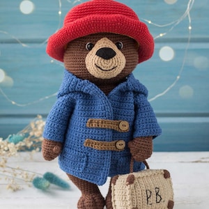 May include: A crocheted brown bear wearing a red hat and a blue coat with brown toggles. The bear is holding a small beige suitcase with the initials "P.B." on it. The bear is a handmade item.