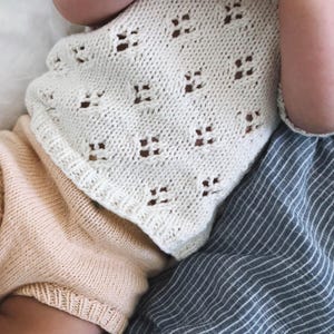 Può includere: Primo piano di una coperta per neonati lavorata a maglia color crema con un motivo floreale. La coperta è ripiegata sugli indumenti di un bambino, tra cui un capo in maglia color pesca e un tessuto a righe blu e bianco.