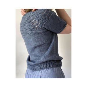 May include: A short-sleeved, slate blue knitted top featuring a decorative lace pattern across the yoke and shoulders. The garment has a ribbed neckline, ribbed sleeve cuffs, and a wide ribbed hem, constructed with a smooth stockinette stitch body.