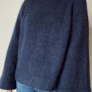 Puede incluir: Un suéter azul marino con cuello barco y mangas largas. El suéter tiene un aspecto suave y texturizado y se combina con unos vaqueros azules. Las mangas son anchas y acampanadas en la muñeca.
