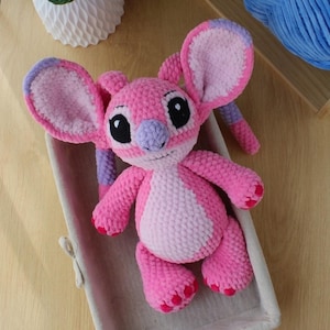 Puede incluir: Un peluche de ganchillo hecho a mano que representa un personaje alien&iacute;gena rosa y rosa claro con orejas grandes, nariz morada y ojos negros. La criatura tiene un acabado de hilo texturizado, extremidades peque&ntilde;as y detalles en rosa.