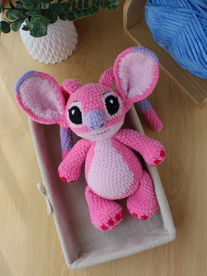 Puede incluir: Un peluche de ganchillo hecho a mano que representa un personaje alien&iacute;gena rosa y rosa claro con orejas grandes, nariz morada y ojos negros. La criatura tiene un acabado de hilo texturizado, extremidades peque&ntilde;as y detalles en rosa.