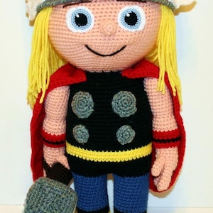 May include: A crocheted doll featuring a character with long yellow yarn hair, a grey helmet with white wings, a black torso with four grey circular accents, a red cape, blue pants, and yellow boots, holding a grey rectangular hammer in one hand.