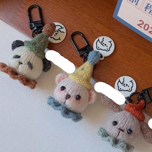 May include: Three handmade keychains featuring knitted animal designs: a panda, a bear, and a bunny. Each keychain has a black clasp and a small round charm with a cartoon face. The panda wears a green hat, the bear a yellow hat, and the bunny an orange hat.
