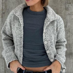 Puede incluir: Un cárdigan de punto jaspeado en gris claro con cuello ancho y mangas largas, que presenta un cierre frontal con botones blancos. Debajo, se lleva una camiseta de canalé en gris carbón oscuro con cuello redondo, creando un estilo superpuesto.