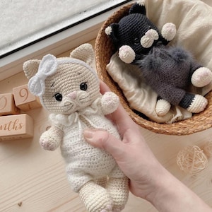 May include: Handmade crochet cat doll in cream with a white bow and a black crochet cat doll in a wicker basket. The cream cat has pink accents on its paws and nose. The black cat has white accents. The dolls are on a light wooden surface.