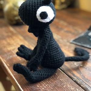May include: A black crocheted stuffed animal featuring a large, round white eye with a black pupil, a small bow tie at the neck, and a long, thin tail. The creature is crafted from black yarn with a visible textured stitch pattern throughout its body.