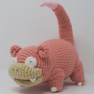 May include: A handmade crochet Slowpoke plush toy in shades of pink and beige. The plush features a pink body, a beige snout, white eyes, and a curled tail with a pink tip. The toy is standing on four small feet with white accents.