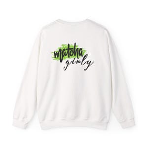 Könnte beinhalten: Weißes Sweatshirt mit Rundhalsausschnitt und dem Text "matcha girly" in schwarzer Schrift über einem grünen Pinselstrich-Design. Das Sweatshirt hat lange Ärmel und eine lockere Passform.