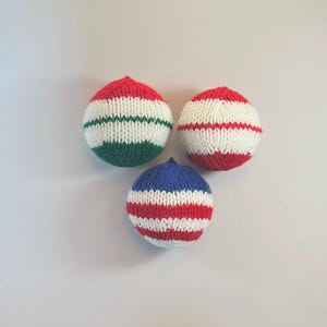 May include: Three hand-knitted balls in red, white, green, and blue. Two balls have red and white stripes, one has red and white stripes, and the third has red, white, and green stripes. The balls are likely for play or decoration.