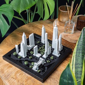 May include: A square black frame containing a white 3D model of a city skyline featuring various skyscrapers and a bridge. The model is set against a black base with small green clusters representing trees, creating a detailed architectural display.