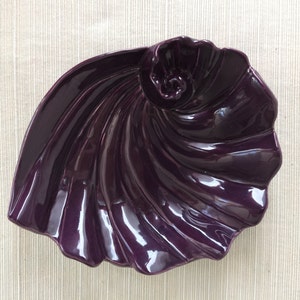 May include: A purple ceramic dish shaped like a seashell. The dish has a glossy finish and a swirling design.
