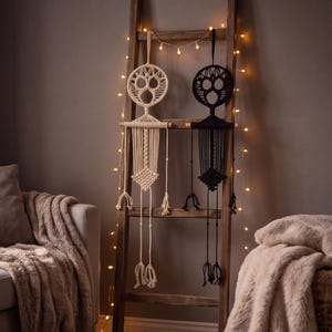 May include: Two macrame wall hangings, one cream and one black, featuring a circular top section with three cutouts, a horizontal wooden dowel, and a long, knotted cord body ending in tassels, suspended from a wooden ladder with string lights.