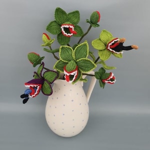 Op de afbeelding: Een grillige opstelling van gehaakte bloemen in een crèmekleurige kan met blauwe stippen. De bloemen zijn groen met rode en witte accenten, sommige hebben open monden met tanden. De stelen zijn bruin en de achtergrond is effen grijs.