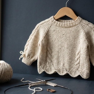 May include: A cream-coloured, speckled knit jumper featuring long sleeves with small side ties, a ribbed crew neckline, and a scalloped hemline. The garment displays intricate cable knit detailing along the front and sleeves, hanging on a wooden hanger.