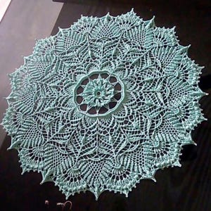 May include: A round, crocheted doily in a soft teal color. The intricate design features a central floral motif surrounded by detailed lace patterns. The edges are scalloped, adding a delicate touch to the handmade textile.