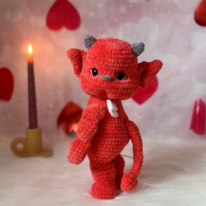 May include: A handmade red crochet devil doll with grey horns and ears, and a small white accent on its chest. The plush toy has black eyes and is standing in front of a candle and heart-shaped decorations.
