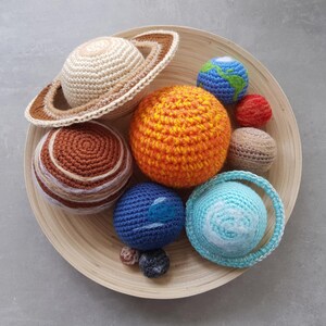Puede incluir: Una colección de nueve esferas de ganchillo que representan planetas y un sol, con texturas y colores variados, incluyendo un gran sol naranja, un planeta beige con anillos, un planeta azul claro con anillos y esferas más pequeñas en varios tonos.