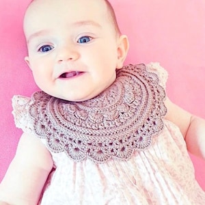 May include: A light brown, round, crocheted collar bib featuring a decorative scalloped edge and intricate stitch patterns. The bib is worn over a light pink sleeveless dress with a subtle white floral print, creating a layered appearance.