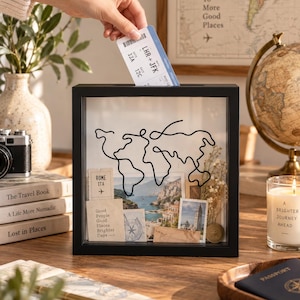 May include: A square black shadow box frame with a glass front featuring a black wire outline of a world map. Inside the frame are travel-themed items including a Rome ITA ticket, a photograph of a coastal town, and a small gold coin.