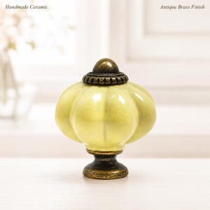 Può includere: Un pomello in ceramica fatto a mano con una finitura giallo pallido lucida e una forma a zucca. La ferramenta presenta una finitura in ottone anticato sulla base e sul tappo superiore. Il testo Handmade Ceramic e Antique Brass Finish è visibile.