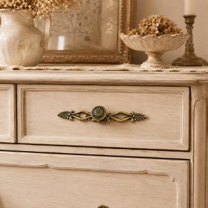 Peut inclure: Un tiroir de commode en bois de couleur crème doté d'une poignée en métal ornée, aux tons laiton antique. La poignée présente un bouton circulaire central avec un motif floral texturé, flanqué de bras décoratifs allongés en volutes.
