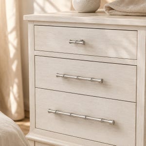 May include: A light-coloured wooden nightstand featuring three drawers with a visible wood grain texture. Each drawer is equipped with a silver-toned metal handle designed to resemble bamboo stalks, providing a metallic contrast to the light finish.
