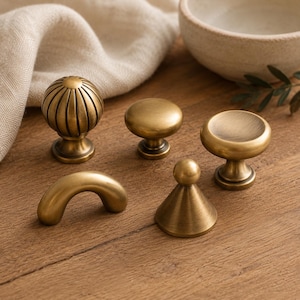 May include: A collection of five brass cabinet knobs and pulls in various shapes, including a fluted sphere, a smooth dome, a wide flat-topped knob, a conical knob with a ball top, and a curved arch-shaped handle, all with a brushed finish.