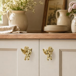 May include: Two gold-colored metal cabinet knobs, each shaped like a small winged cherub figure. The knobs are mounted on light-colored cabinet doors, featuring a polished, reflective surface that catches the light, highlighting the detailed form.