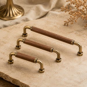 Peut inclure: Trois poignées de placard de longueurs différentes, chacune dotée d'une poignée cylindrique en bois brun foncé centrée entre deux montants métalliques incurvés à finition laiton antique avec des bases de fixation circulaires.