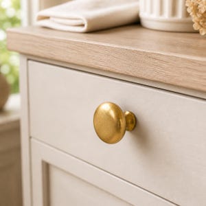 May include: A round, brushed gold cabinet knob with a smooth, domed surface and a cylindrical base, mounted on a light gray drawer front. The knob features a metallic finish that reflects light, contrasting with the neutral-toned furniture piece.