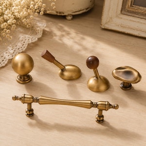 May include: A collection of brass hardware including a long horizontal handle with decorative ends, a spherical knob, a mushroom-shaped knob, and two wall hooks featuring a combination of brass bases and dark brown wooden accents.