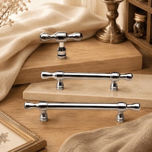 May include: Three polished chrome cabinet pulls with a cylindrical bar design and rounded ends. The set includes one T-shaped knob and two longer handles, each featuring a smooth, reflective metallic finish and tiered circular mounting bases.