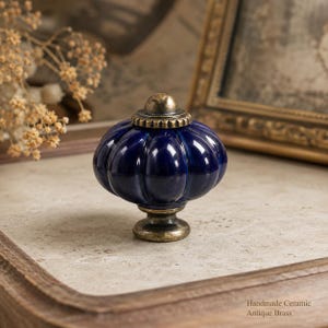 Può includere: Un pomello per mobili in ceramica blu scuro con corpo a forma di zucca e base e tappo superiore con finitura in ottone antico. Il pomello presenta una superficie liscia e lucida con creste verticali e il testo Handmade Ceramic Antique Brass.
