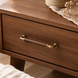 Puede incluir: Un cajón de madera marrón oscuro con un tirador horizontal compuesto por un cilindro central de madera oscura, flanqueado por dos extremos y soportes de montaje metálicos en tono latón, fijados al panel frontal del mueble.