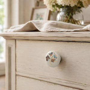 May include: A round white ceramic drawer knob featuring a delicate hand-painted botanical design of small brown and blue leaves with thin golden stems, attached to a light beige wooden drawer front with a visible wood grain texture.