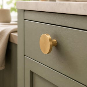 May include: A circular, brushed brass cabinet knob with a flat face and a cylindrical base, mounted on a sage green wooden drawer front. The knob features a smooth, metallic finish and a simple, geometric design that protrudes from the drawer.