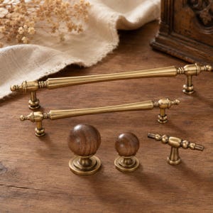 May include: A collection of cabinet hardware including two long brass drawer pulls with tapered centers, two round wooden knobs of different sizes, and one small brass T-bar handle. The hardware features a metallic gold finish and natural wood grain.