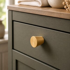 May include: A matte gold, conical-shaped cabinet knob with a flat circular face, mounted on a dark olive green drawer front. The knob features a smooth, brushed metallic finish and a tapered body that extends outward from the drawer surface.